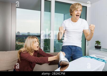 Cheerful man cheering while shocked woman at the ironing board in living room Stock Photo
