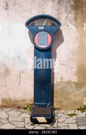 A vintage blue public weighing scale stands on a stone-paved ground against a weathered, textured wall. The scale features a circular dial with a red background and a printed chart, and a metal Stock Photo