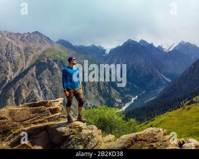 The traveler s guy stands on a rock in the background of a beautiful mountain valley. A guy in a blue jacket on a background of a mountain gorge. The Stock Photo