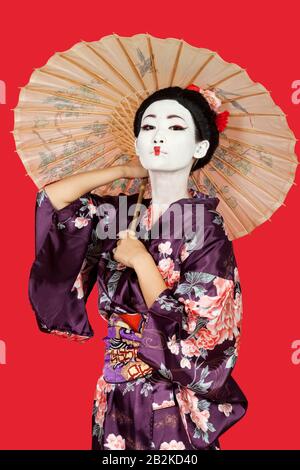 Portrait of Japanese woman in kimono holding parasol against red background Stock Photo