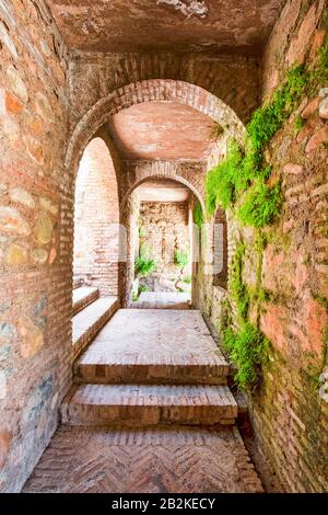 A narrow, covered passageway constructed from aged red brick and stone features a series of rounded arches leading into the distance. The floor is paved with herringbone-patterned bricks, and a set Stock Photo