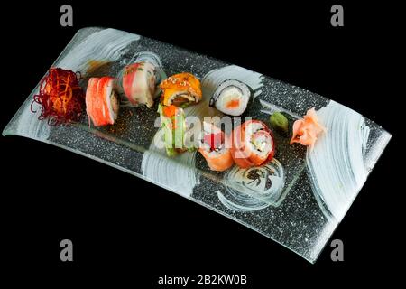 A variety of sushi rolls are arranged on a rectangular, textured glass plate with a black and white abstract design. The sushi includes different types such as those wrapped in salmon, avocado, crab Stock Photo