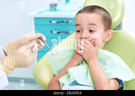 Portrait of a scared child in a dental chair. The boy covers his mouth with his hands, afraid of being examined by a dentist. Children's dentistry. Stock Photo