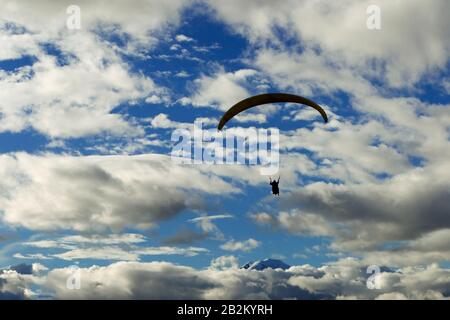 A paraglider is seen in silhouette against a vibrant blue sky filled with scattered white and gray clouds. The paraglider's canopy is arched and dark, with the pilot suspended below, holding control Stock Photo