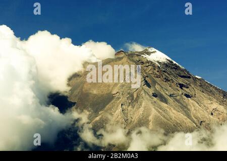 A rugged mountain peak with snow on its summit rises above a thick layer of white clouds. The rocky slopes are exposed, showing layers of earthy tones and textures, with some dark rock outcrops. The Stock Photo