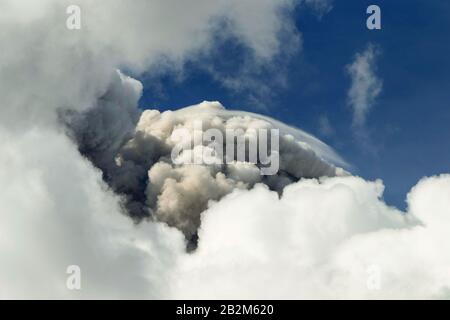 A large, dense plume of smoke, appearing gray and white, rises vertically into the atmosphere, forming a towering, billowing column against a deep blue sky. The smoke is surrounded by white cumulus Stock Photo