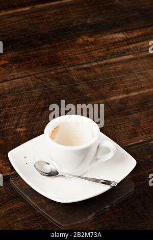 A white ceramic coffee cup, partially empty with coffee residue inside, sits on a matching white square saucer. A small metal spoon rests on the saucer beside the cup. The set is placed on a dark Stock Photo