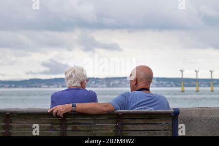Back view of man and woman sat on a wooden bench overlooking Portsmouth harbour on a cloudy summer day. Stock Photo