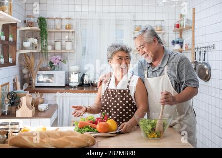 Senior couple having fun in kitchen with healthy food - Retired people cooking meal at home with man and woman preparing lunch with bio vegetables Stock Photo