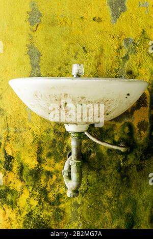 A weathered white ceramic sink is mounted on a textured yellow wall covered in patches of green moss and peeling paint. The sink shows significant grime, rust stains, and discoloration, with a single Stock Photo