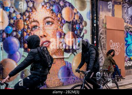 Two hooded youth cycle past a huge piece of graffiti art while a lady sits nearby and looks at the camera. Stock Photo