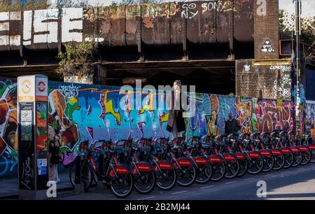 A young Asian man stands on a bicycle in the middle of a row of parked bikes for hire, to have his photo taken in Brick Lane, London. Stock Photo