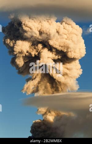 A large, dense plume of smoke billows upward into a clear blue sky. The smoke appears thick and turbulent, with varying shades from dark gray to light beige, suggesting intense heat or combustion Stock Photo