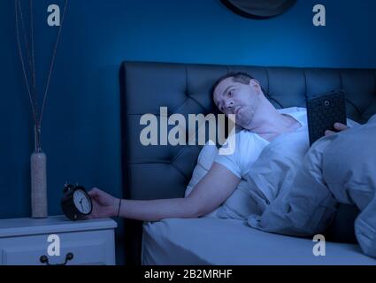 a man working on his tablet at night looks at his alarm clock and cannot sleep Stock Photo