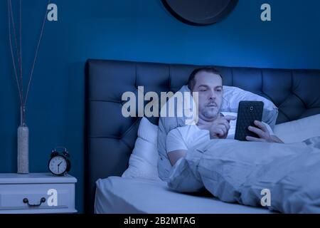 a man is lying in bed playing on the tablet at night and cannot sleep Stock Photo