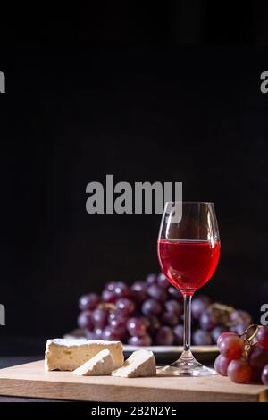 A glass of red fruit wine. Close-up of grape vine and brie cheese on a dark background in low key. Copy space. Banner for cafe, winery and restaurant. Stock Photo