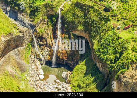 An aerial perspective shows a waterfall plunging from a high cliff into a circular pool below, surrounded by rugged rock formations and lush green vegetation. The cliff face features exposed earth Stock Photo