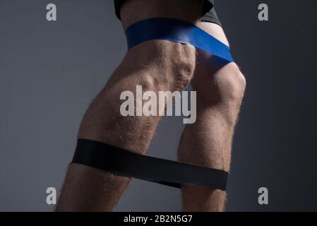 closeup, bottom view of the guys muscular legs in sports shorts, stretching fitness elastic bands with his legs, involvement of leg muscle groups that Stock Photo