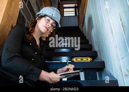 low angle view of female inspector sitting on the stair, house stairs review as she taking professional notes using a clipboard and looking to camera Stock Photo