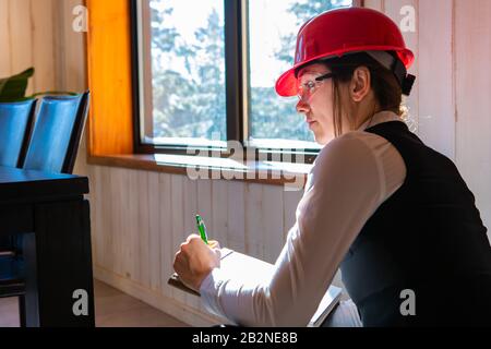 construction inspector during pre-purchase inspection of a home interior. young caucasian woman wearing red hard hat using the clipboard to take notes Stock Photo