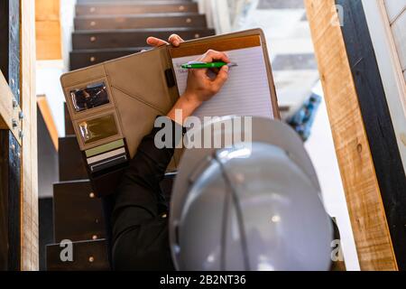 top view of construction inspector woman sits on the stairs during a home pre-purchase inspection, she takes professional notes on notebook clipboard Stock Photo