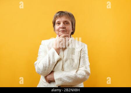 Portrait of a pensive thinking gray-haired senior woman holding her chin with her hand on a yellow background.