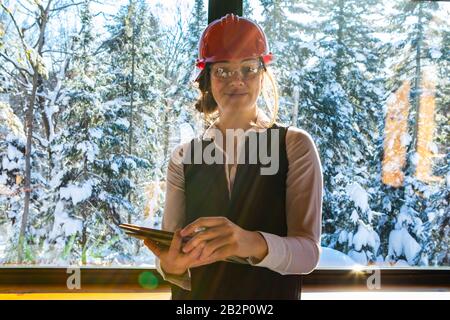 Woman architect standing interior on the front of a glass window, with sun rays and snowy trees outdoor as she smiling and holding a clipboard.  Stock Photo