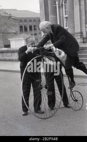 1980s, historical, an elderly man attempting to get on or mount a penny-farthing bicycle, also known as a high wheeler or an 'ordinary', York, England. The machine was the first to be known as a 'bicycle' and with its large front wheel was capable of high speeds. However it was difficult to mount, ride and there was a danger of falling of and it was superceded by the modern 'safety' bicycle. Stock Photo