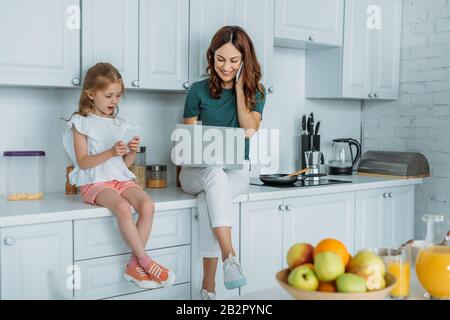 woman talking on smartphone and using laptop while sitting on kitchen counter near daughter using smartphone Stock Photo