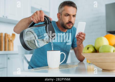 selective focus of man overfilling cup with water while sitting at kitchen table and using smartphone Stock Photo