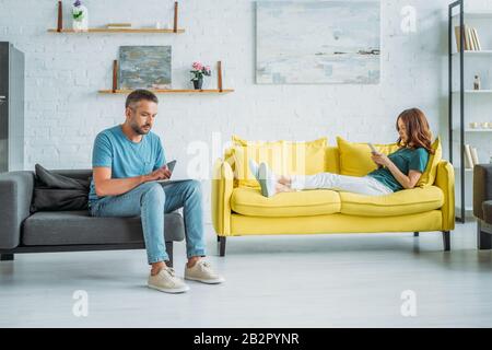 woman lying on yellow sofa with smartphone near husband sitting on couch and using smartphone Stock Photo