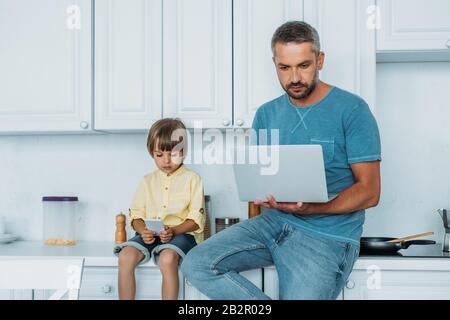 father using laptop and and son using smartphone while sitting on kitchen counter together Stock Photo