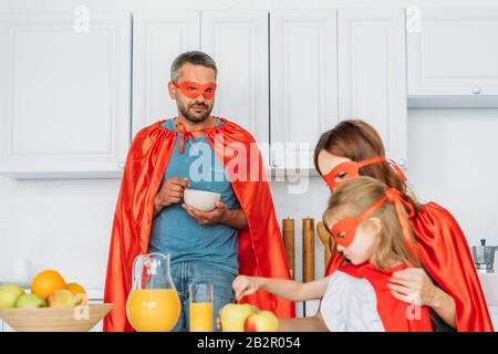 father, mother and daughter in costumes of superheroes having breakfast in kitchen Stock Photo