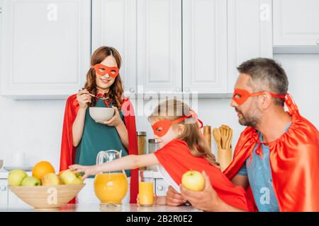 family in costumes of superheroes having breakfast in kitchen together Stock Photo