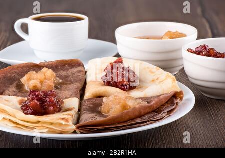 Pancakes are plain and with cocoa. Pancakes on a white plate, apple jam of different varieties and coffee over a wooden background. Traditional Russia Stock Photo