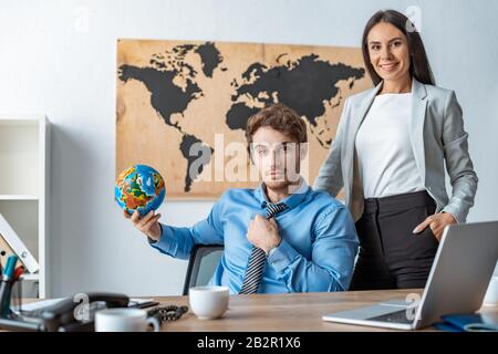 attractive travel agent standing with hand in pocket near colleague holding globe Stock Photo