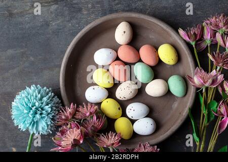 Easter eggs painted in pastel colors on a vintage wooden background texture, concrete look plate surrounded with various spring flowers in modern Stock Photo