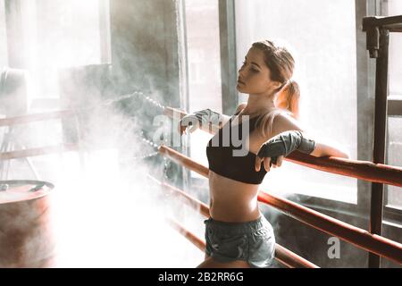 Young pretty woman standing on ring and resting. Close-up portrait of female fit girl at a boxing studio with bright light in the background. Stock Photo
