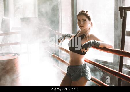 Young pretty woman standing on ring and resting. Close-up portrait of female fit girl at a boxing studio with bright light in the background. Stock Photo