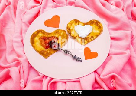 Heart shaped pancakes for romantic breakfast with strawberry jam, silver spoon and paper hearts. Shrovetide (carnival) concept. On pink cloth linen ba Stock Photo