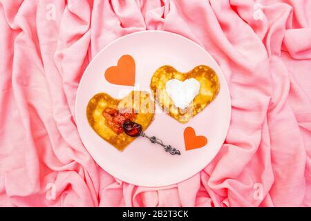 Heart shaped pancakes for romantic breakfast with strawberry jam, silver spoon and paper hearts. Shrovetide (carnival) concept. On pink cloth linen ba Stock Photo