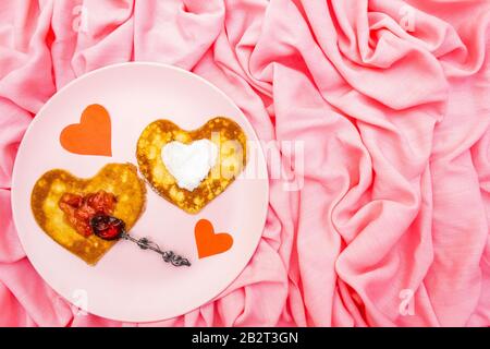 Heart shaped pancakes for romantic breakfast with strawberry jam, silver spoon and paper hearts. Shrovetide (carnival) concept. On pink cloth linen ba Stock Photo