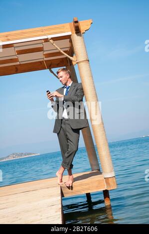 Barefoot businessman relaxing with his smartphone standing outdoors on a waterfront dock Stock Photo