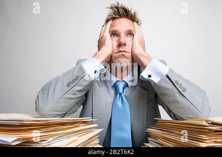 Bored businessman holding his head in his hands on the piles of paperwork on his desk Stock Photo