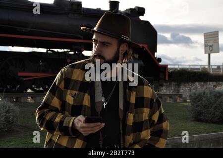 Bearded man wearing a cowboy hat, next to a train station using a smartphone/ Young man wearing a 1950 fedora hat Stock Photo