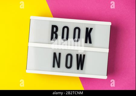 Book now - text on a display lightbox on yellow and pink background. Stock Photo