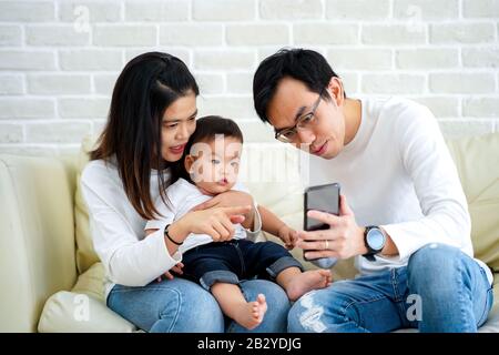 Cheerful young Asian family with son watching video on mobile phone and sitting on sofa couch at home, parents and kid enjoying cartoon movie Stock Photo