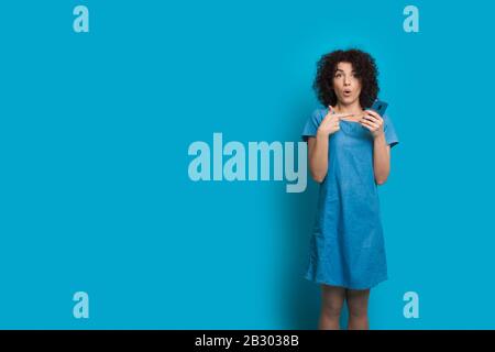 Shocked caucasian girl with curly hair is pointing to her mobile phone while posing on blue wall with blank space Stock Photo