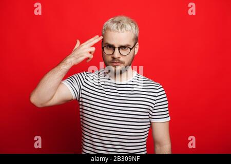 Serious blonde bearded man looking through eyeglasses and gesturing like shooting in head while posing on red background Stock Photo