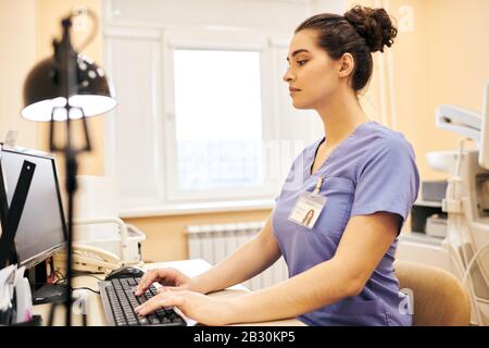 Concentrated young medical clinic employee in scrubs sitting at table and typing on computer keyboard Stock Photo